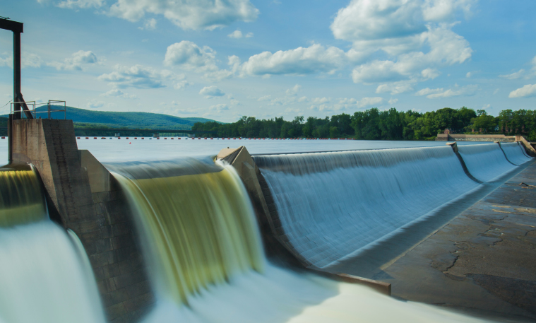 Water flows over a dam in a scenic setting.