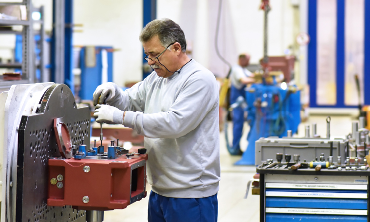 Man wearing a white sweatshirt, blue jeans, and gloves works on repairing a gearbox in a factory that houses industrial equipment.