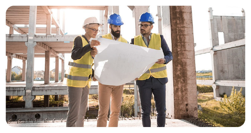 Three architects on a construction site look at building plans while wearing safety gear.