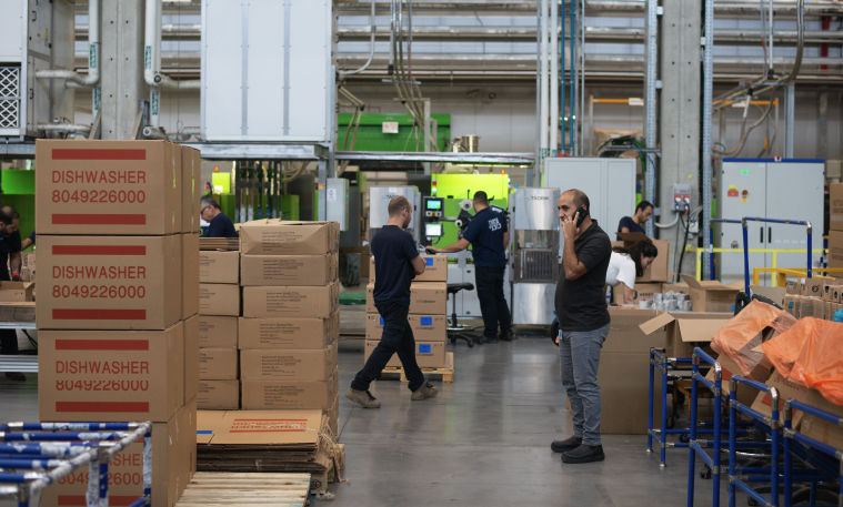 A large warehouse is packed with boxes containing dishwashers as employees work on machinery and the foreman makes a call on a cellphone.